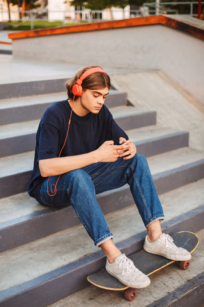 Joven skater en auriculares naranjas usando cuidadosamente el teléfono celular sentado en las escaleras con skate clásico en el skate park
 - Foto, imagen