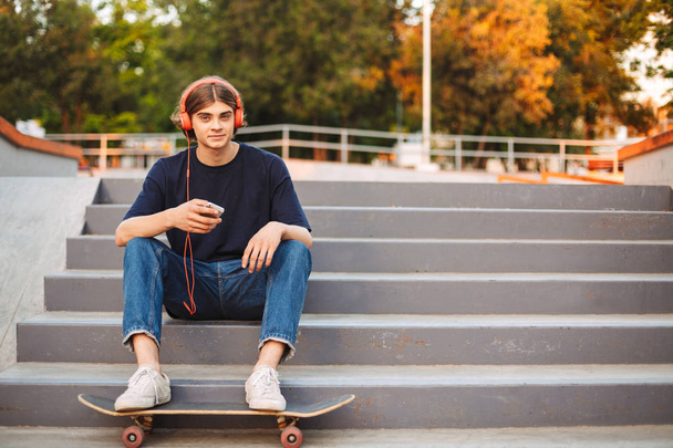 Young skater in orange headphones holding cellphone in hands happily looking in camera on stairs with skateboard at skate park - Photo, Image