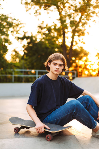Jovem patinador em camiseta preta e jeans olhando atenciosamente na câmera com skate passando tempo no parque de skate
 - Foto, Imagem