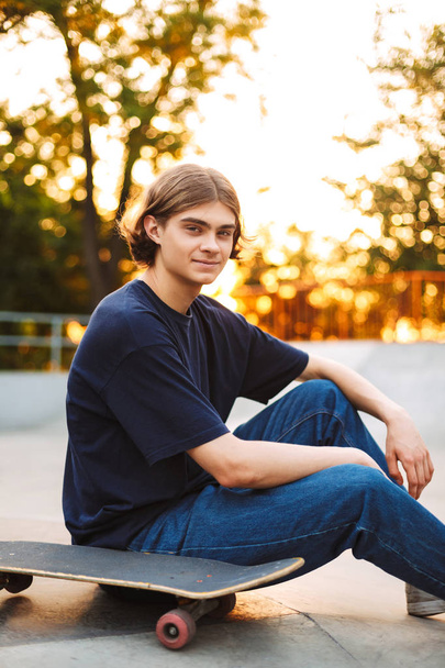 junger lächelnder Skater in schwarzem T-Shirt und Jeans, der verträumt in die Kamera schaut und Zeit mit Skateboard im modernen Skatepark verbringt  - Foto, Bild