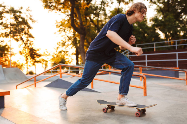 Νέοι δροσερό σκέιτερ σε μαύρο T-shirt και τζιν ιππασία στην skateskateboard στο πάρκο skate με την Ανατολή του ηλίου σε φόντο - Φωτογραφία, εικόνα