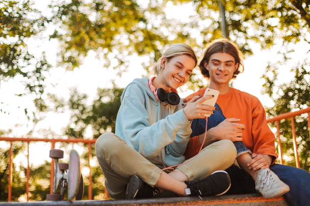 Giovane ragazzo sorridente e ragazza felicemente utilizzando il cellulare trascorrere del tempo insieme a skatepark
  - Foto, immagini