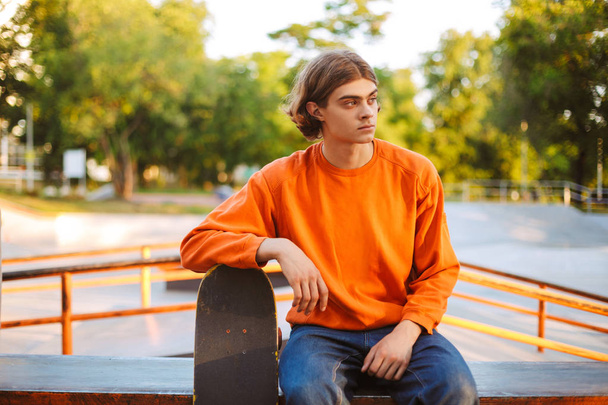 Молодой фигурист в оранжевом пуловере задумчиво смотрит в сторону, опираясь на скейтборд с современным скейтпарком на заднем плане
 - Фото, изображение