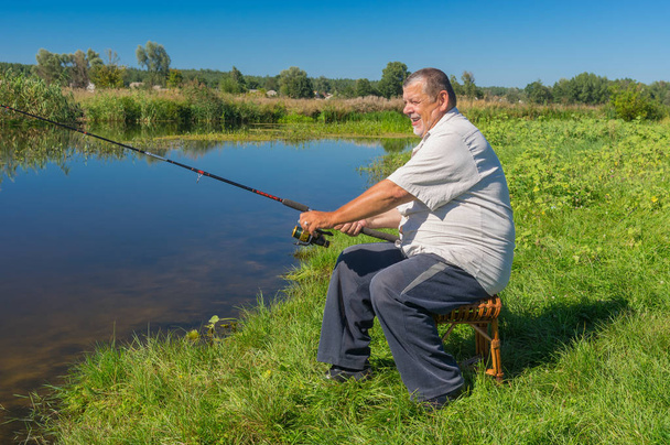 Pescador sênior sentado em um banquinho de vime com vara giratória e pronto para pescar no pequeno rio Merla, no centro da Ucrânia
 - Foto, Imagem