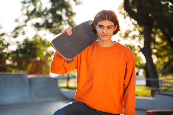 Νεαρός χαμογελαστό σκέιτερ στον πορτοκαλί πουλόβερ ευτυχώς ψάχνει στην κάμερα skateboard εκμετάλλευση στον ώμο με skatepark στο φόντο - Φωτογραφία, εικόνα