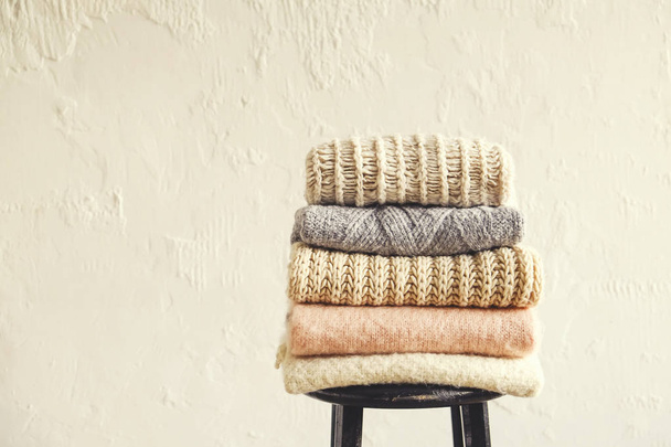 Bunch of knitted pastel color sweaters, different knitting patterns folded in neat stack on black wooden barstool, grunged texture wall background. Fall winter season knitwear. Close up, copy space. - 写真・画像