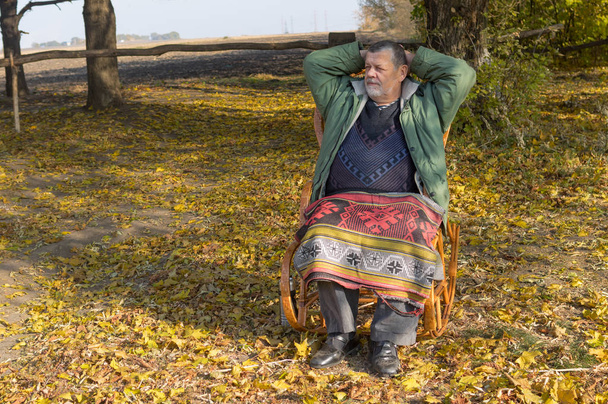 Ανώτερος άνθρωπος κάθεται σε ψάθινη καρέκλα στην ηλιόλουστη φθινοπωρινή μέρα - Φωτογραφία, εικόνα