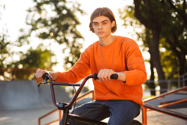 Nadenkende jongen in oranje trui zittend op fiets dromerig tijd in de camera doorbrengen kijken naar moderne skatepark - Foto, afbeelding