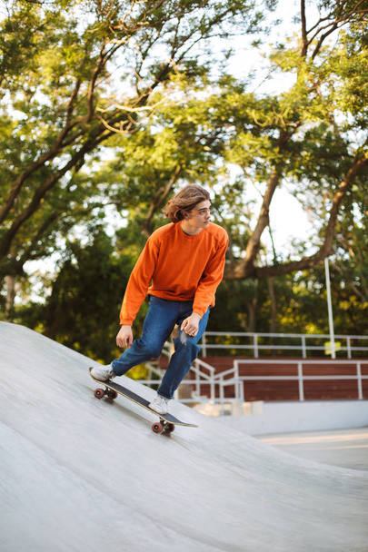 Ο νεαρός άντρας σε πορτοκαλί πουλόβερ skateboard και εν ενεργεία ακροβατικά που αφιερώνουν χρόνο στο σύγχρονο skatepark - Φωτογραφία, εικόνα