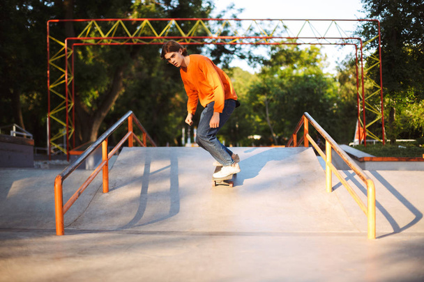 Молодой вдумчивый скейтбордист в оранжевом пуловере и практикующий трюки, проводящий время в современном скейтпарке
 - Фото, изображение