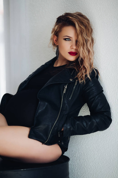 Gorgeous pregnant woman in leather jacket and professional make-up leaning on white background. - Foto, afbeelding