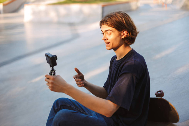 Young smiling guy happily recording new video for vlog with skateboard near at modern skatepark - Photo, Image