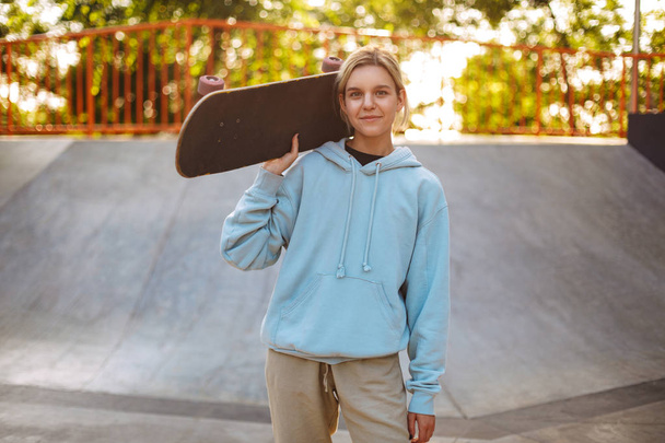 Pretty smiling girl in hoodie holding skateboard on shoulder dreamily looking in camera at skatepark - Photo, Image