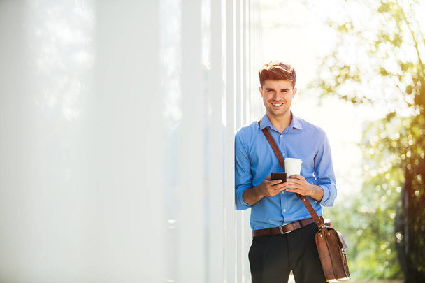 knappe jongeman gaan werken, op kantoor, in blauw shirt met koffie in handen op een zonnige dag - Foto, afbeelding