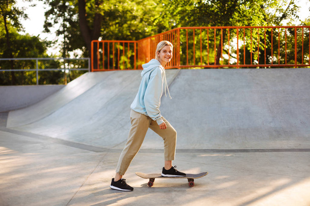 Vrij lachende meisje in hoodie gelukkig kijken in de camera probeert skateboarden in moderne skatepark - Foto, afbeelding