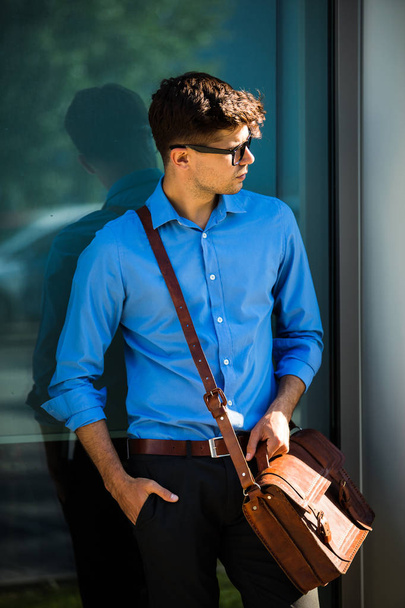 handsome office man with glasses and leather bag, standing against a wall and presenting a side of free space around him - Фото, изображение