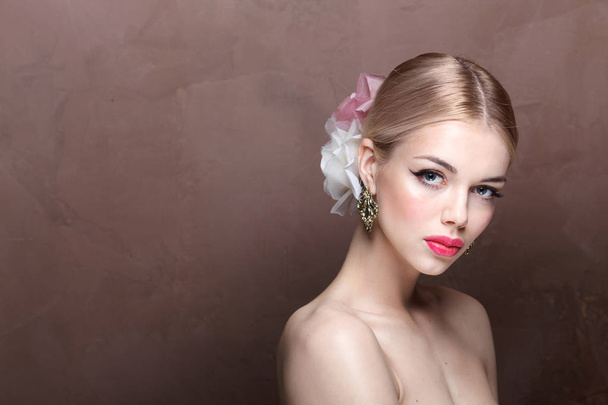 young blonde woman with pink lips and roses in her hair wearing vintage earrings posing on brown background with copy space - Photo, Image