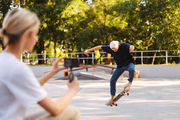 Mladí bruslař cvičí kousky na skateboardu zároveň hezká dívka nahrávání nové video k vlog na skatepark  - Fotografie, Obrázek