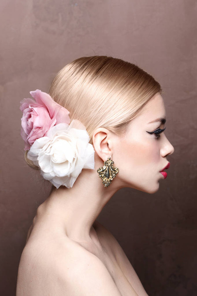 Fashion portrait of young blonde model with pink lips and roses in her hair wearing vintage earrings, side view - Photo, Image