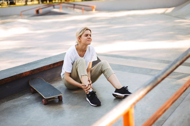 Upset girl sadly closing eyes holding her painful leg with skateboard near at skatepark - Photo, image