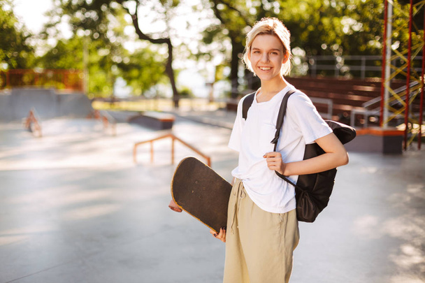 Pretty smiling girl with backpack and skateboard in hand joyfully looking in camera with big modern skatepark on background - Photo, Image