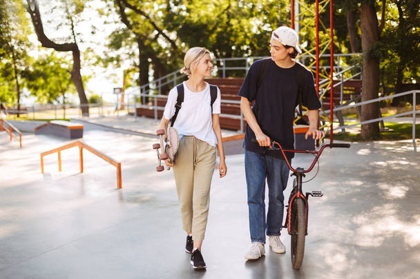 Young guy with bicycle and pretty girl with skateboard happily spending time together at modern skatepark - Фото, изображение