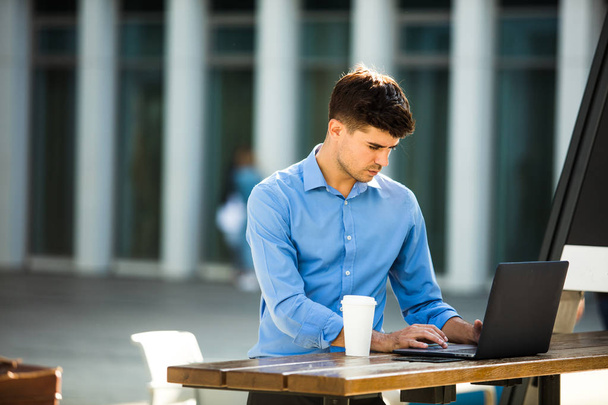enfocado joven hombre de oficina que trabaja desde el exterior en el ordenador portátil con café a su lado, trabajo remoto
 - Foto, imagen