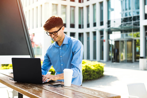 enfocado joven hombre de oficina que trabaja desde el exterior en el ordenador portátil con café a su lado, trabajo remoto
 - Foto, Imagen