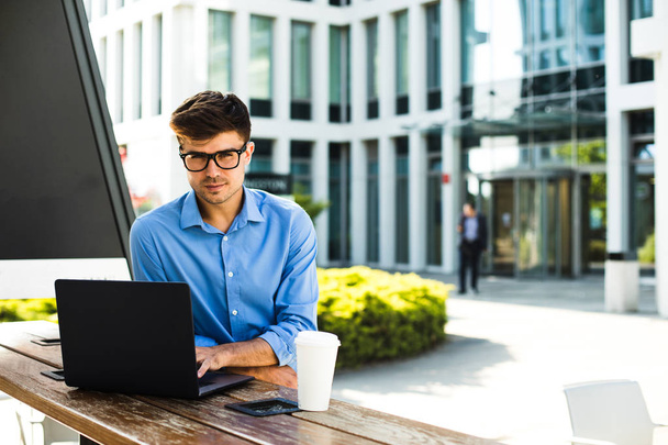 уверенный и улыбающийся бизнесмен, предприимчивый, смотрящий в камеру и работающий снаружи за ноутбуком, имеющий офисное здание за спиной
 - Фото, изображение