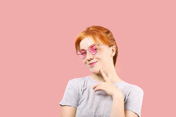 Attractive young woman with long red hair, wearing pink cat eye sunglasses, showing flirtatious body language, touching face. Beautiful redhead female flirty facial expression. Background, copy space. - Zdjęcie, obraz