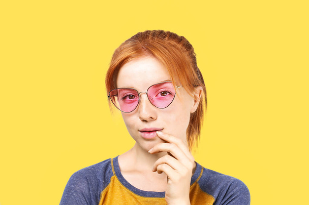 Attractive young woman with long red hair, wearing pink cat eye sunglasses, showing flirtatious body language, touching face. Beautiful redhead female flirty facial expression. Background, copy space. - Фото, зображення