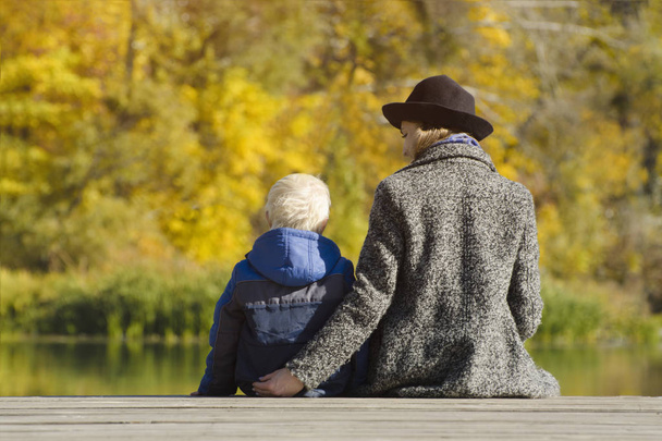 金髪の少年と彼の母親は、ドックの上に座って。秋の日。背面図 - 写真・画像
