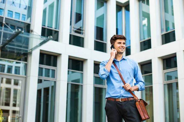 confident and successful antrepreneur, office man or corporate guy, standing with his leather bag and elegant outfit in front of an office building and talking at cellphone - Фото, зображення