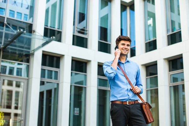confident and successful antrepreneur, office man or corporate guy, standing with his leather bag and elegant outfit in front of an office building and talking at cellphone - Zdjęcie, obraz