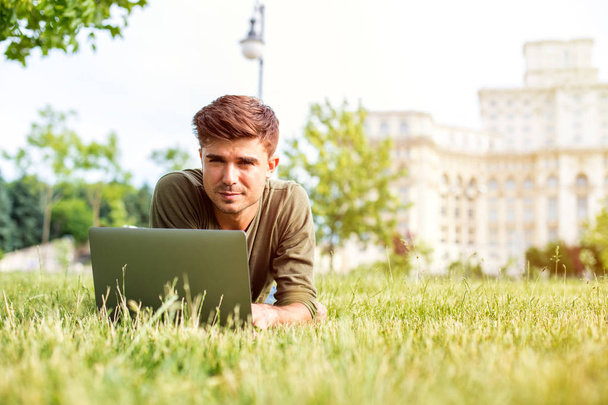 handsome young man, student, lying in grass, looking or working on laptop, outside in park in center of a city - Photo, Image