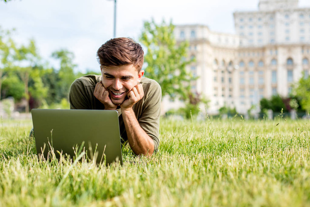 handsome young man, student, lying in grass, looking or working on laptop, outside in park in center of a city - Foto, immagini