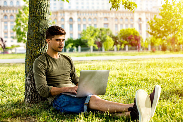 handsome young man, student, lying in grass against a tree, looking or working on laptop, outside in park in center of a city - Valokuva, kuva