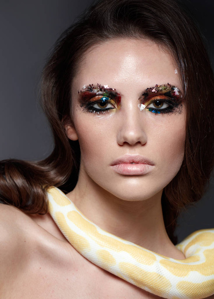 fashion portrait of brunette model with color spangles on eyelids and yellow snake lying on her neck - Fotografie, Obrázek