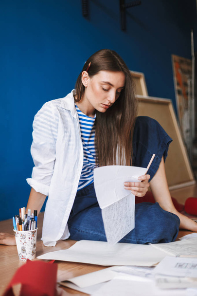 Задумчивая девушка в белой рубашке и полосатой футболке сидит на полу держа в руке лист бумаги с холстами на заднем плане дома
 - Фото, изображение