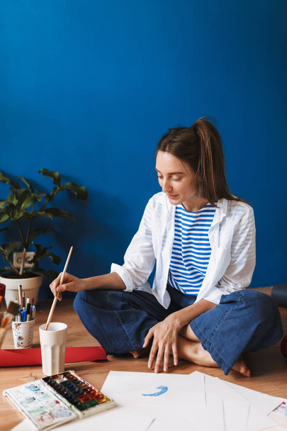Young smiling painter in white shirt and striped T-shirt sitting on floor dreamily drawing spending time at cozy home - Foto, immagini