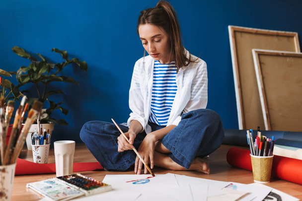 Pensive girl in white shirt and striped T-shirt sitting on floor dreamily drawing by watercolor spending time at home - Foto, Imagen