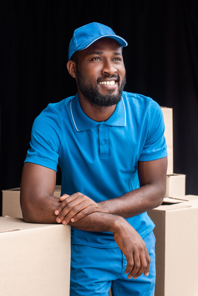 smiling african american delivery man leaning on boxes and looking away - 写真・画像