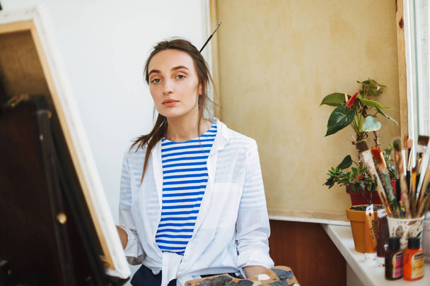Beautiful girl in white shirt and striped T-shirt dreamily looking in camera drawing on easel with paint tools near on window sill at home - Photo, Image