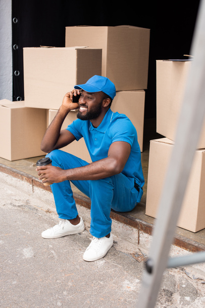 african american delivery man talking by smartphone and holding coffee in paper cup - 写真・画像