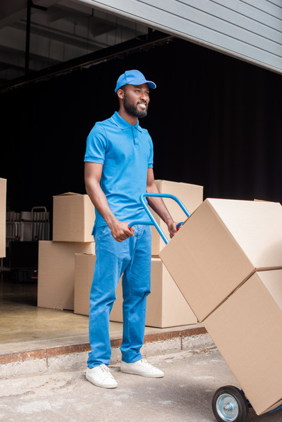 smiling african american delivery man holding cart with boxes and looking away - Photo, Image