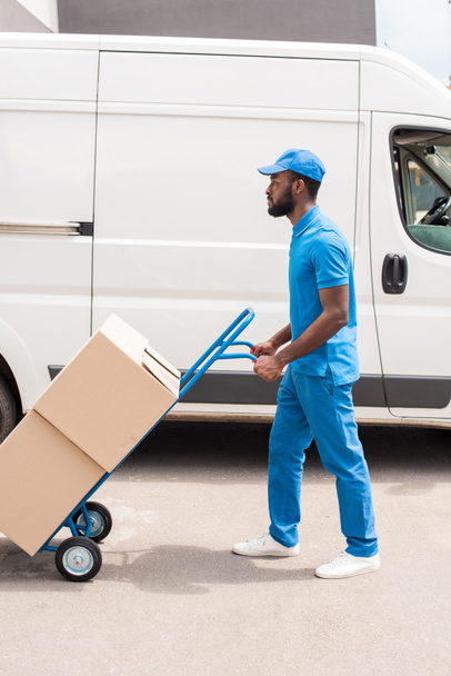 side view of african american delivery man with cart and boxes near van - 写真・画像