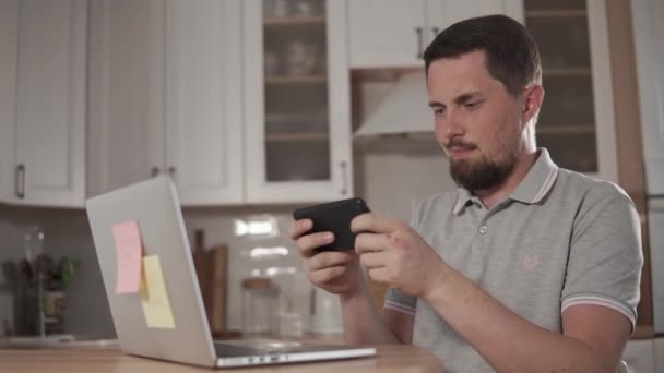 Adult handsome man is playing on a smartphone in home, wearing headphones - Footage, Video