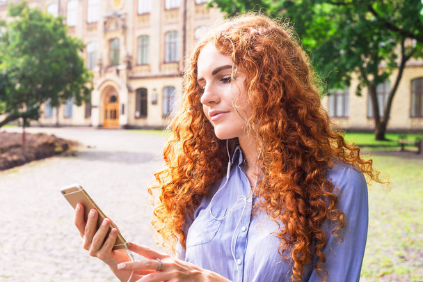 Uma linda garota com cabelos ruivos encaracolados olha para seu smartphone, ao qual os fones de ouvido estão conectados. Usando um smartphone e tecnologia moderna
 - Foto, Imagem