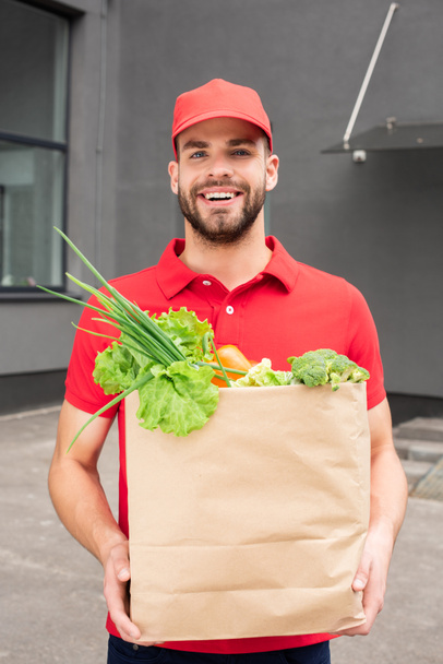 smiling caucasian delivery man holding paper bag with vegetables - Foto, Imagem