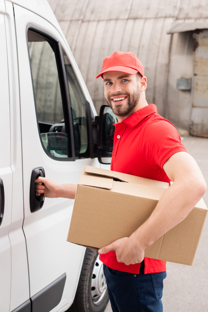 smiling delivery man in uniform with cardboard box standing near white van in street - Foto, Bild